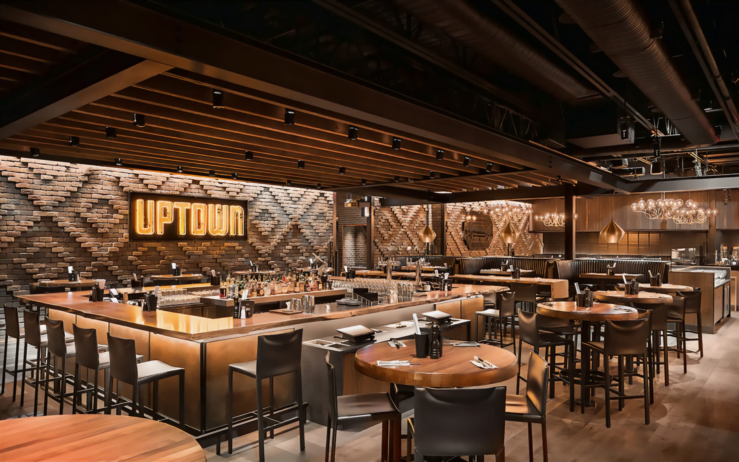 Restaurant interior with ambient lighting, brick walls, high-top tables, black leather booths, and a bar with a neon 'Uptown' sign behind it.