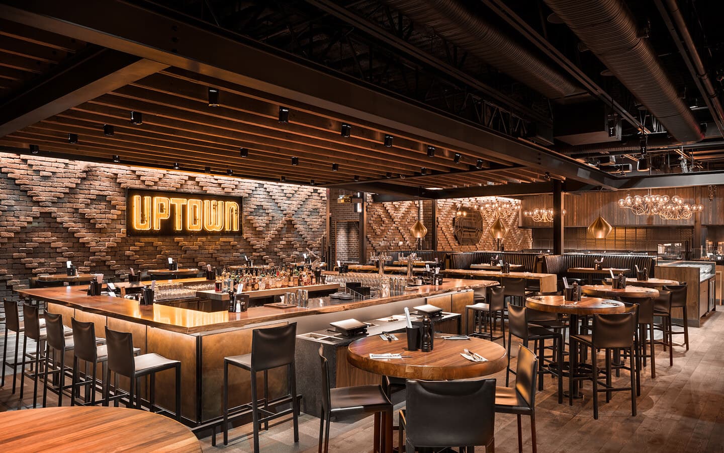 Restaurant interior with ambient lighting, brick walls, high-top tables, black leather booths, and a bar with a neon 'Uptown' sign behind it.