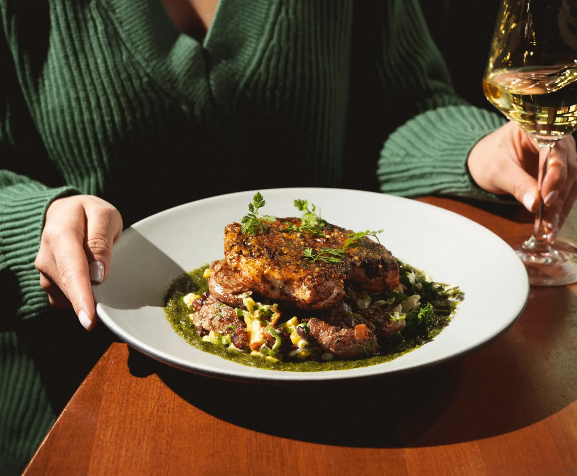 Woman in a green sweater holding a glass of white wine and the Cajun blackened chicken dish on a wooden table.